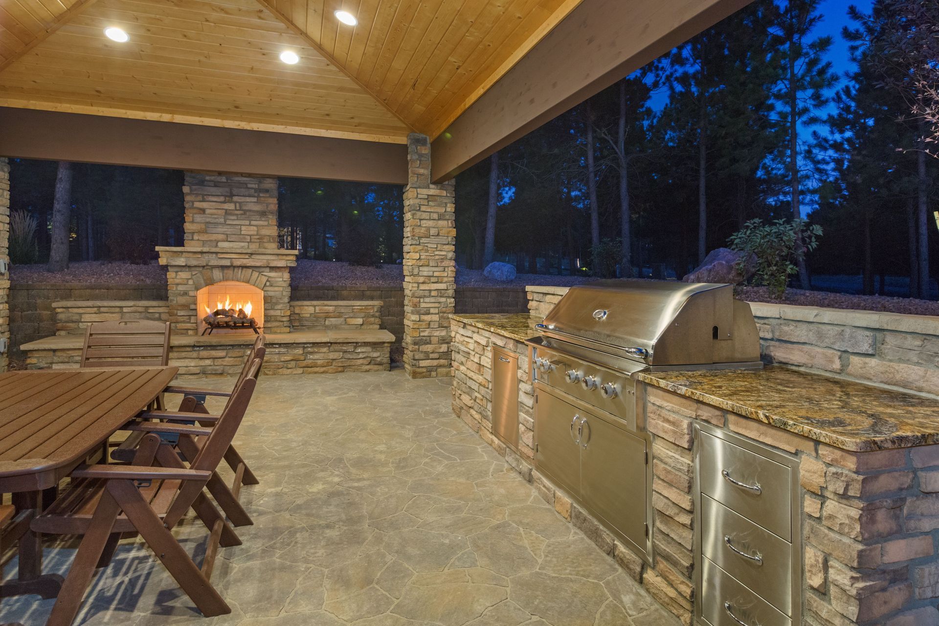 Covered outdoor kitchen with stone grill, built-in counter, picnic table, and warm wood ceiling at dusk