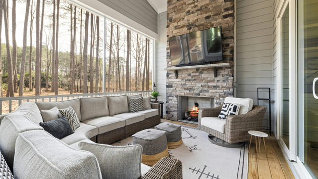 Cozy screened porch with gray sectional, wicker chairs, ottoman, fireplace, and forest view