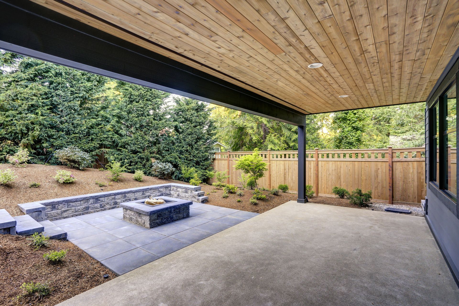 Covered patio with wood ceiling, concrete floor, and sunken fire pit beside a fenced yard