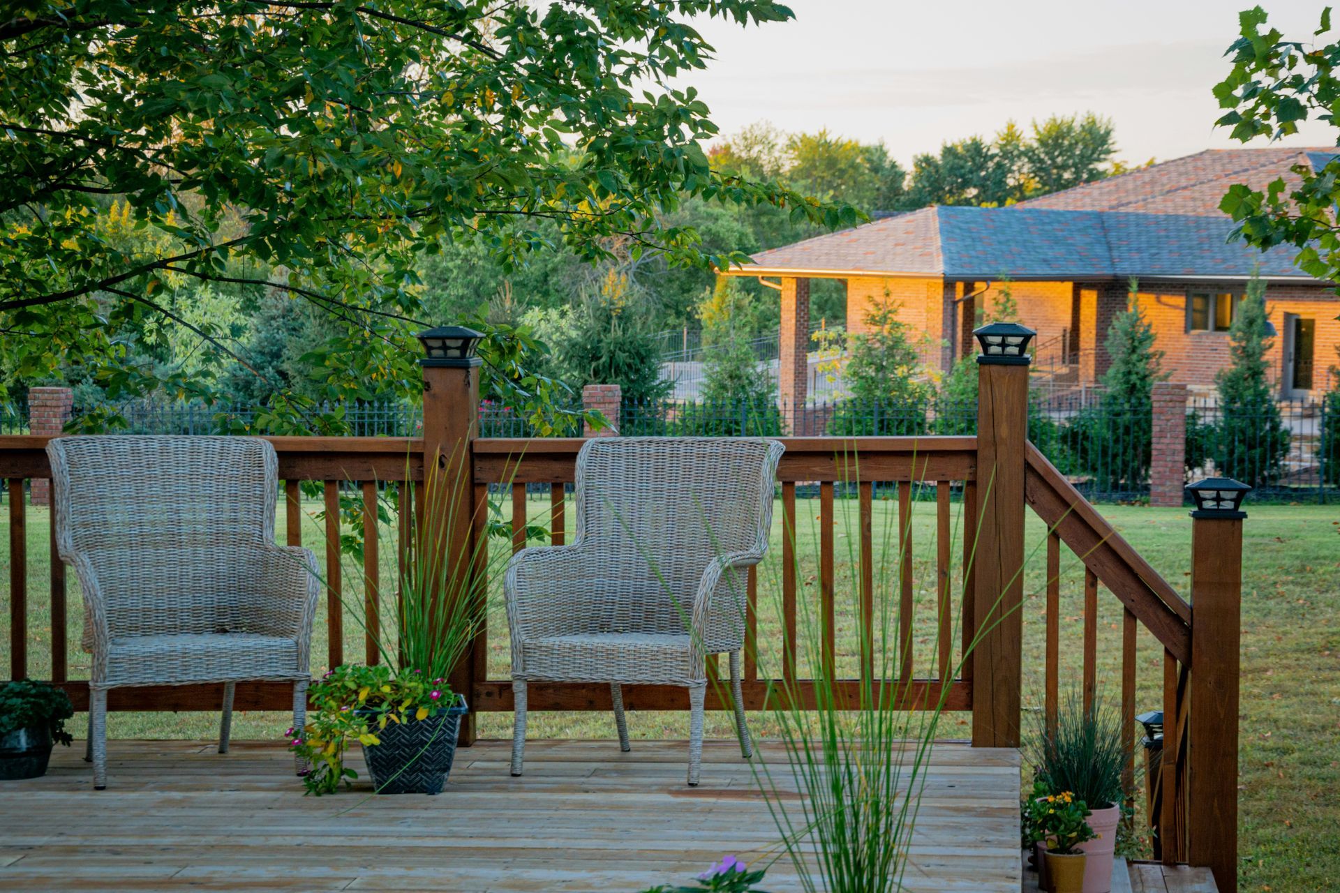 Wooden deck with two wicker chairs, potted plants, and a view of a house and green yard at sunset