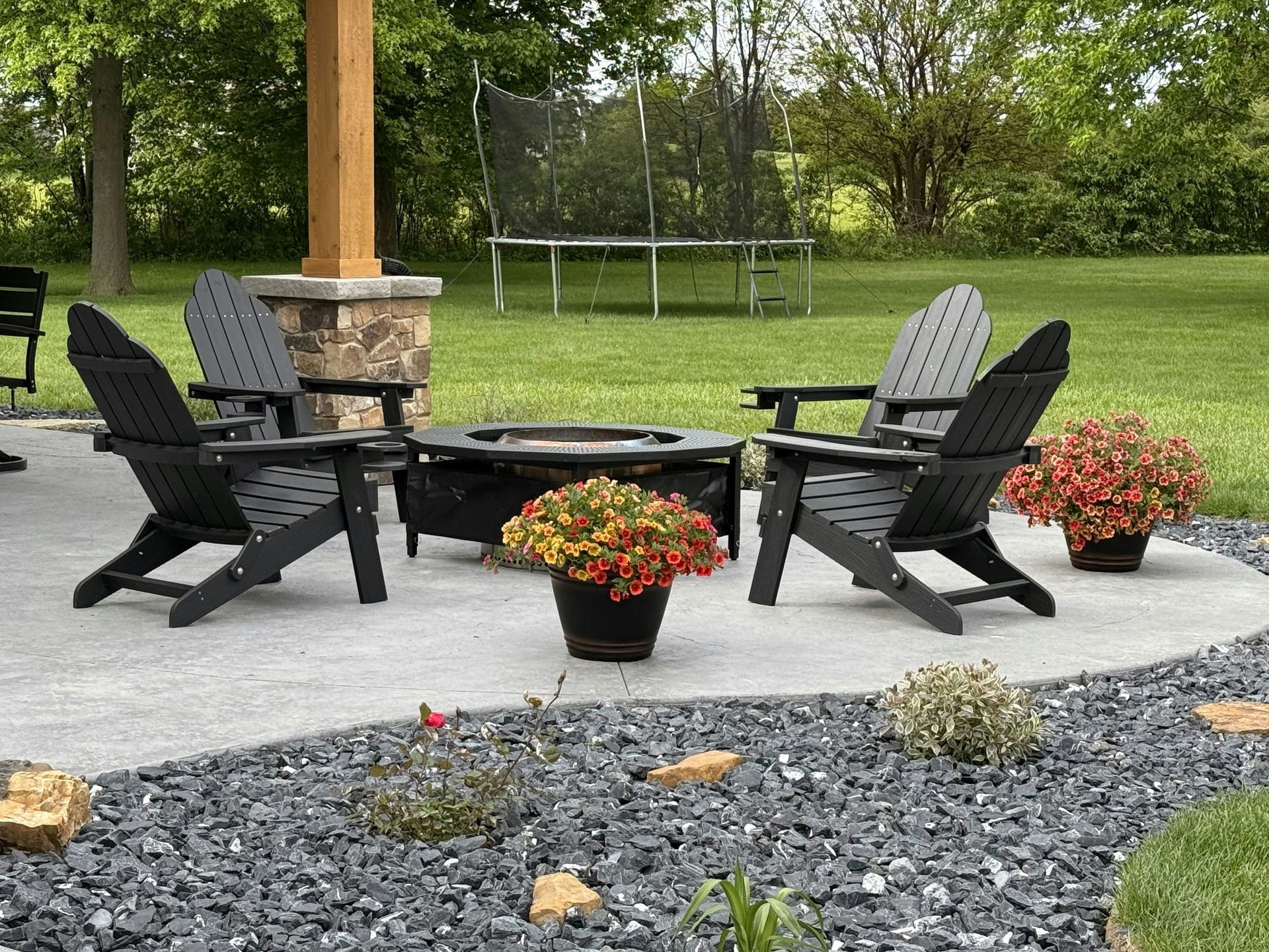 Patio with black chairs around a fire pit, potted flowers, and a grassy yard with a trampoline.