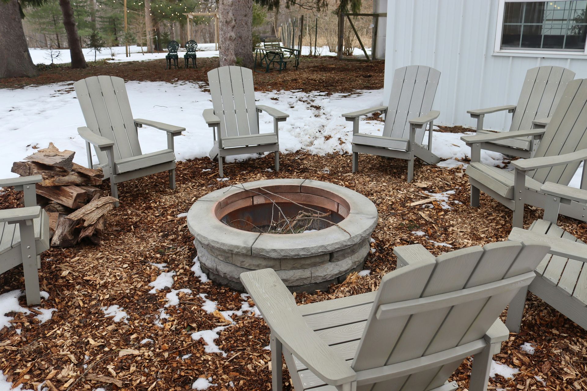 Snowy backyard fire pit surrounded by white Adirondack chairs and woodpiles