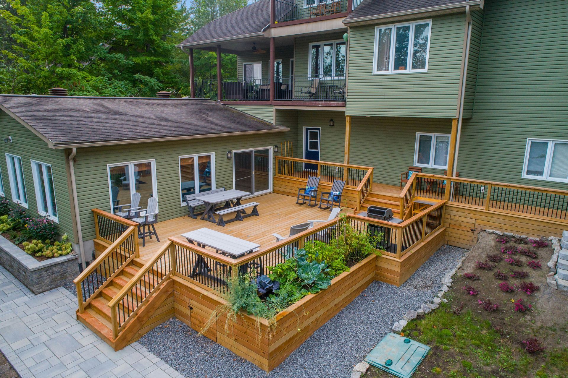 Wooden deck patio with tables, chairs, and planter boxes beside a green house with stairs and railings