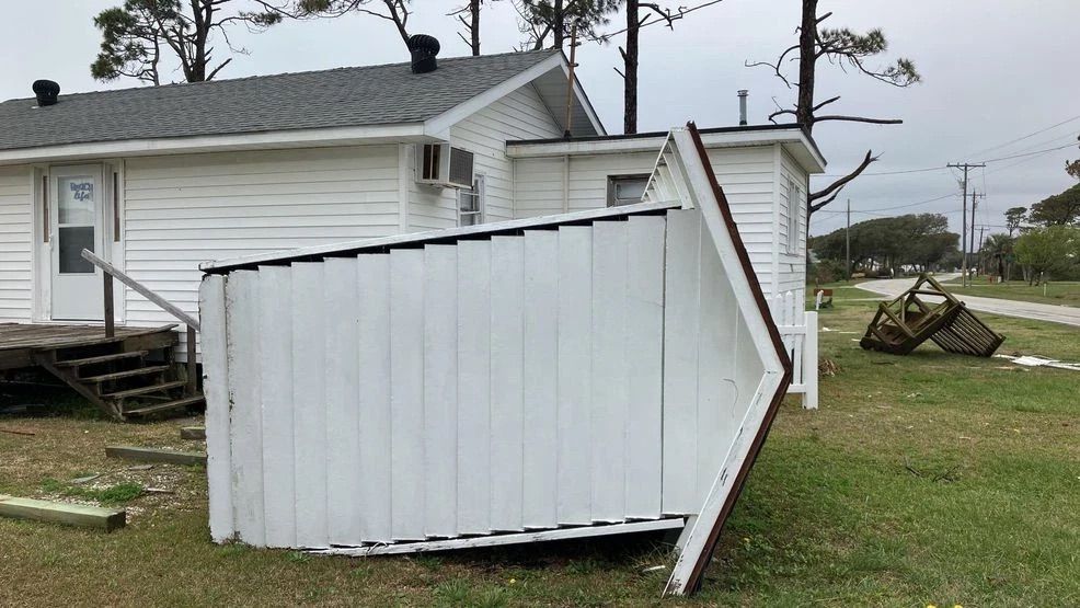 White shed lying on its side in a grassy yard beside a house after storm damage