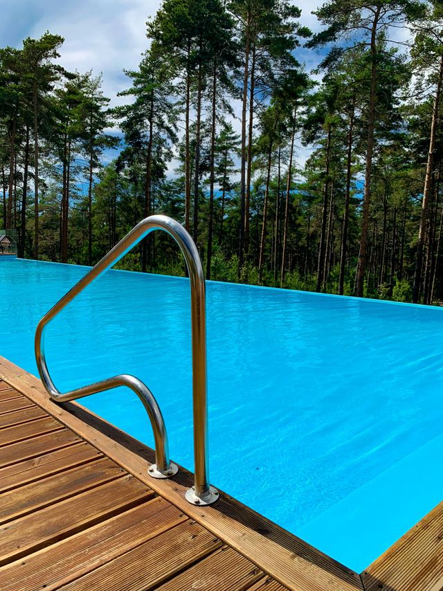 Blue infinity pool with metal ladder beside a wooden deck and trees in the background