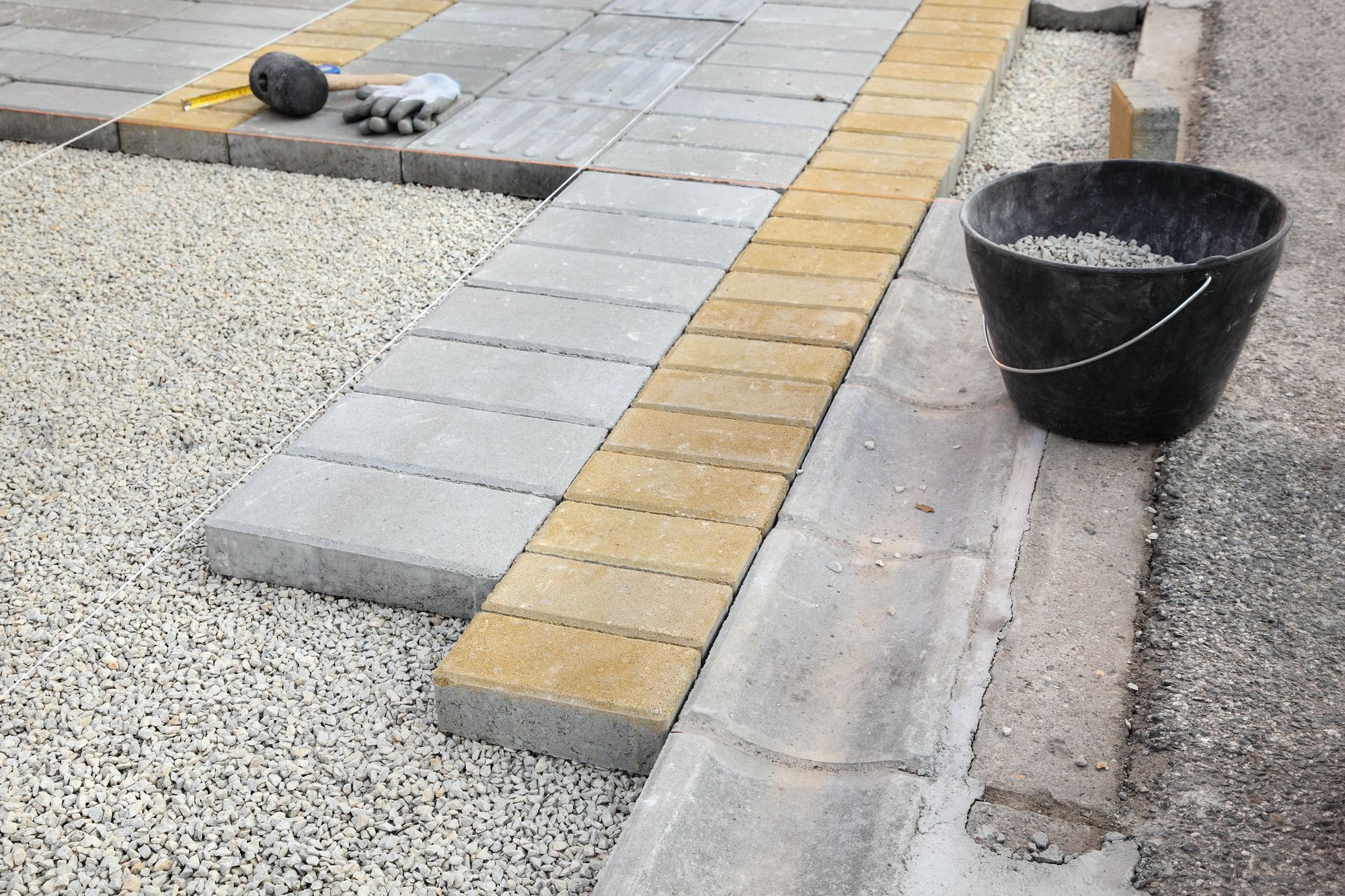 Paved walkway with gray and yellow pavers beside gravel, a curb, and a black bucket of wet cement.