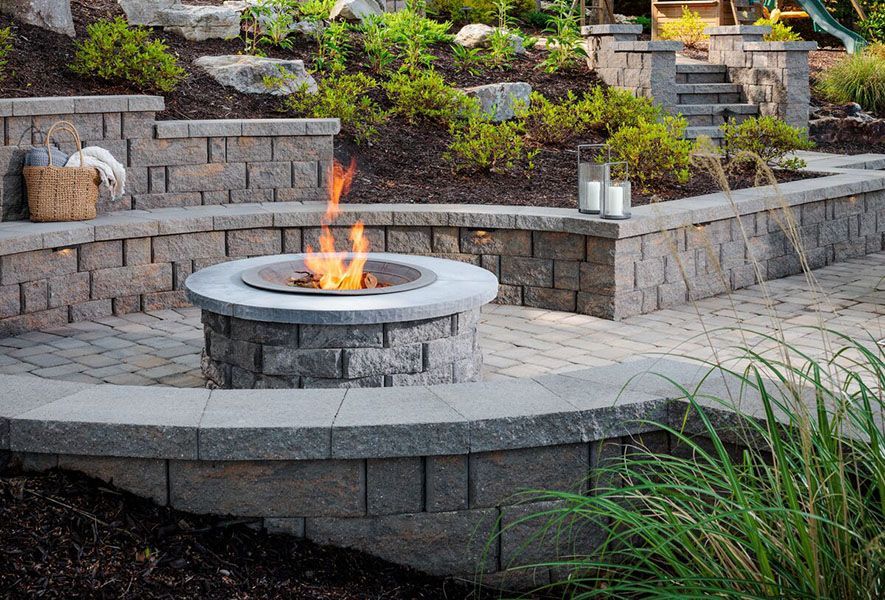 Stone patio fire pit with a small flame, surrounded by tiered retaining walls and landscaping.