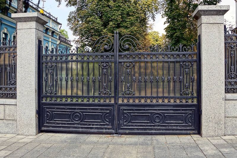 Ornate black iron gate between stone pillars, closed at the entrance to a tree-lined building courtyard