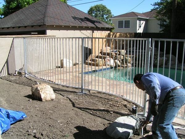 Worker installing a fence around a backyard pool area, with a house and rocks nearby