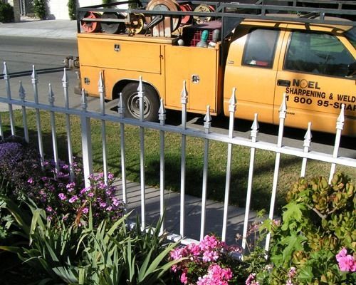 Yellow utility truck parked behind a white fence beside a garden of pink flowers