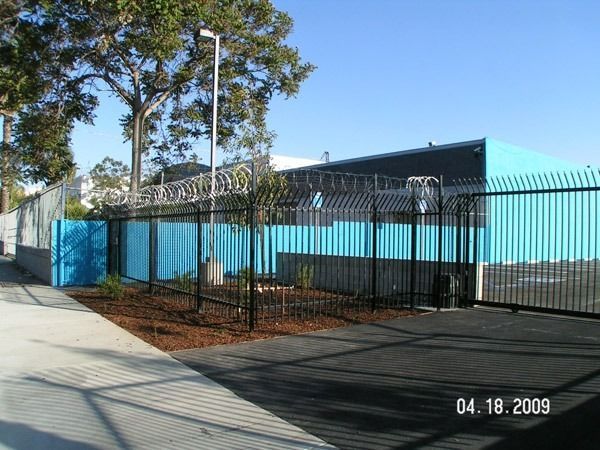 Industrial building behind black fence with blue panels on a sunny street corner