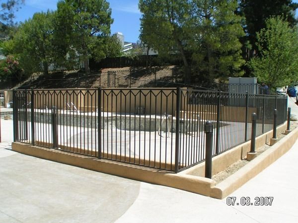 Black metal fence around a raised concrete area with trees and a sidewalk nearby.
