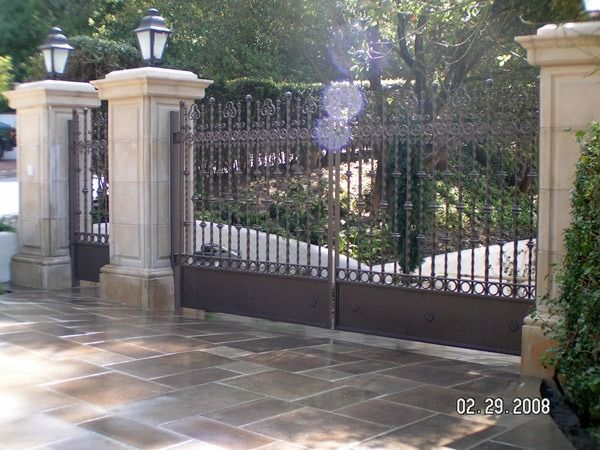 Ornate black wrought-iron gate between stone pillars, with greenery behind and tiled pavement in front.