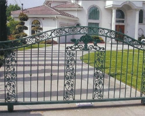 Ornate black metal driveway gate in front of a large white house with a lawn and steps