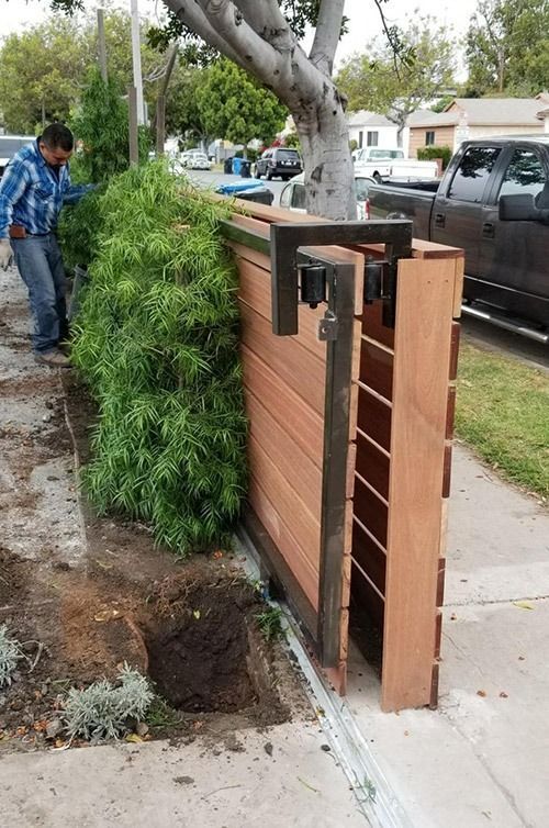 Wooden fence panel leaning beside a sidewalk, with a worker by a fresh hole and parked vehicles nearby.