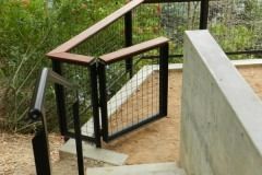 Outdoor stair landing with black metal gate, wooden railing, and concrete wall beside a dirt path