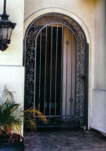 Arched wrought-iron front door with decorative frame beside a wall lantern and potted plant