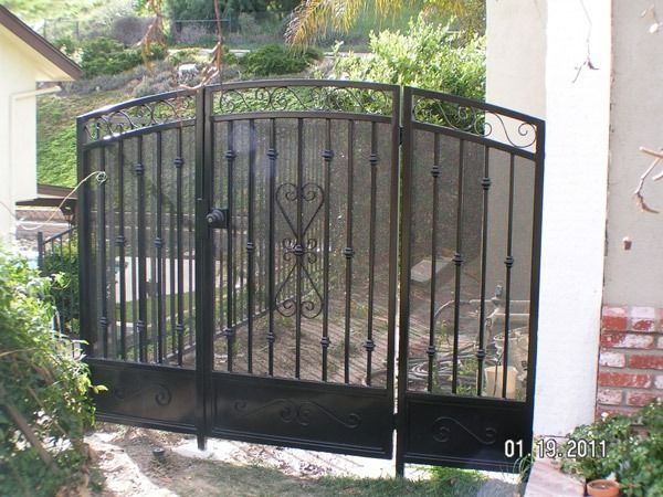 Black metal driveway gate at the entrance of a home, with stone path and garden plants nearby
