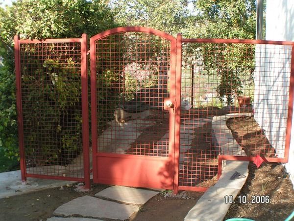 Red metal gate and fenced walkway leading into a leafy yard, with a white wall on the right.