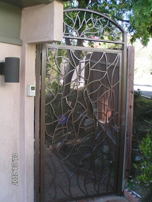 Decorative metal gate with leaf pattern at a house entrance, beside a wall-mounted light and doorbell.