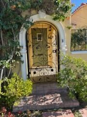 Arched front door with white wrought-iron gate, framed by greenery and a small brick path.