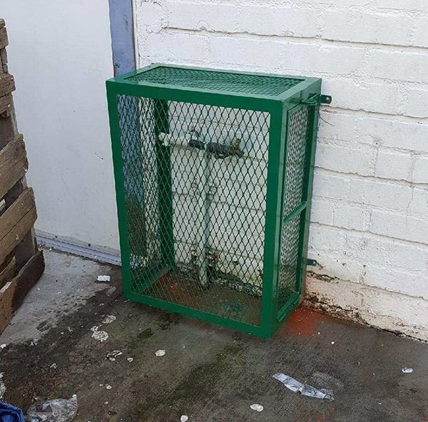 Green metal utility cage with mesh sides, mounted against a white brick wall outdoors.