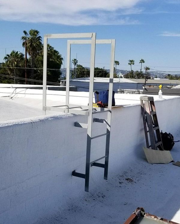 White rooftop access ladder beside a ladder frame on a bright rooftop with palm trees in the distance