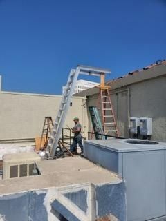 Worker on rooftop beside ladders and HVAC units under a clear blue sky.