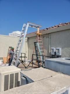 Workers on a rooftop beside ladders and HVAC units, with a blue sky overhead.