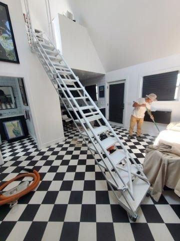 Two people on a ladder in a black-and-white tiled room with a high ceiling, adjusting a window blind