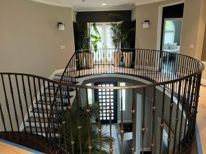 Curved indoor staircase with black railings overlooking a bright entryway and potted plants.