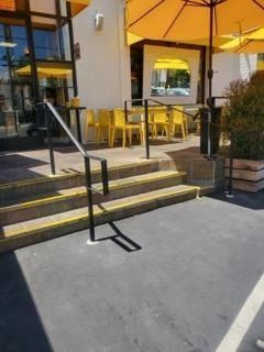 Exterior entrance with steps, black handrails, and a yellow patio seating area by glass doors.
