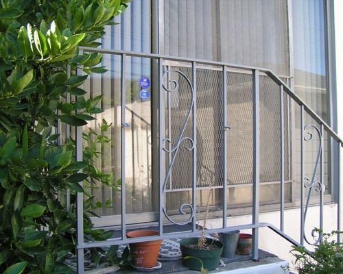 Front porch with metal railing, potted plants, and a mesh security door beside a leafy tree.