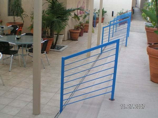 Blue metal railing along a tiled outdoor walkway with potted plants and chairs on the left.