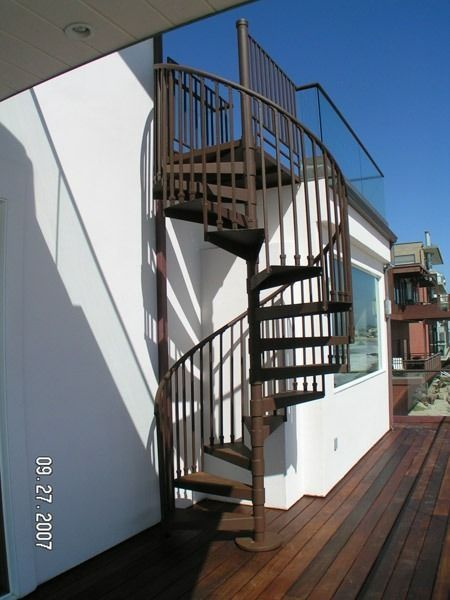 Outdoor metal spiral staircase on a sunlit wooden deck beside a white building