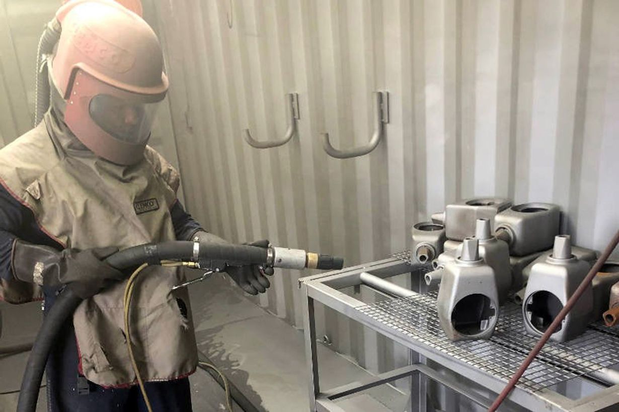 Worker in protective gear spray-painting equipment in a small industrial booth.