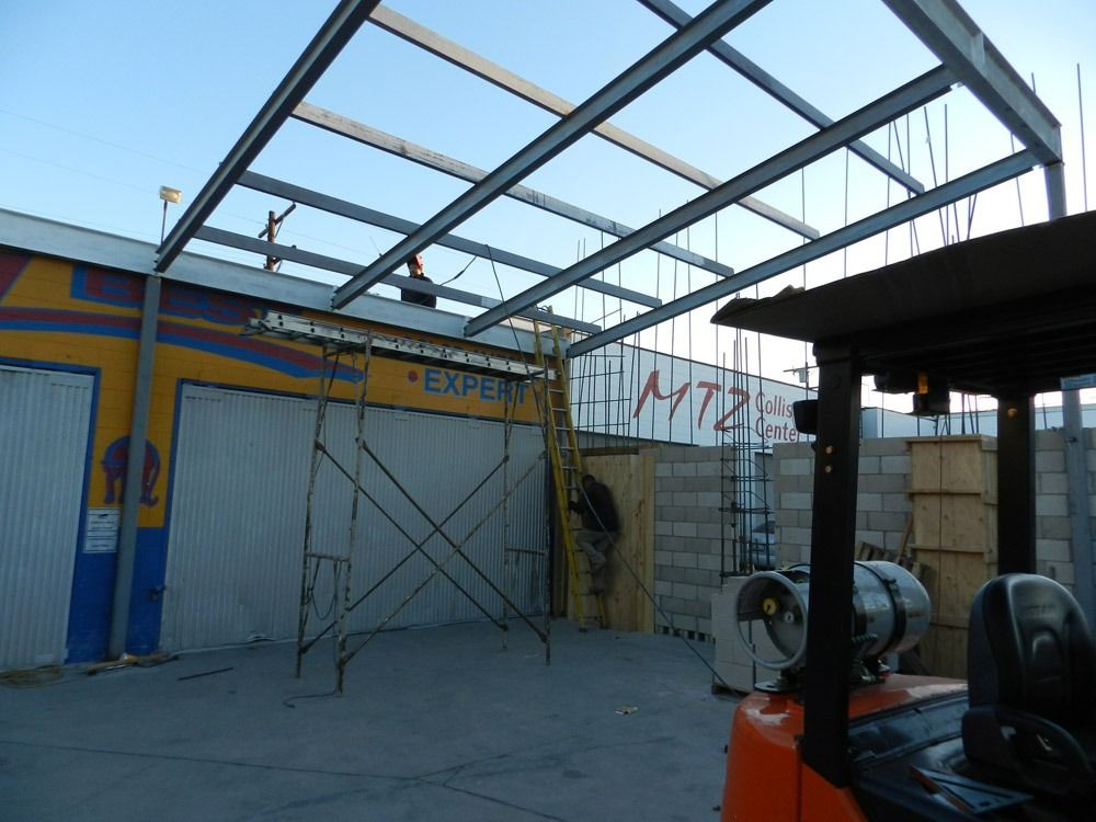 Partially built metal roof frame over a courtyard beside a white wall and parked equipment