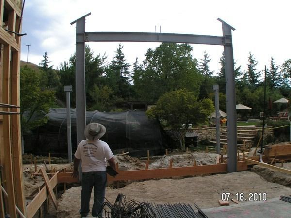 Person standing at a construction site beneath a large steel frame, with trees and materials in the background.