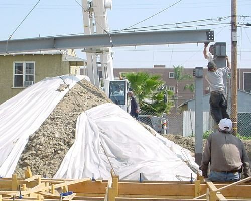 Workers on a construction site lifting a concrete beam over a covered pile of materials
