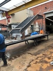 Industrial conveyor belt on a metal frame in a workshop yard, with a worker beside it.