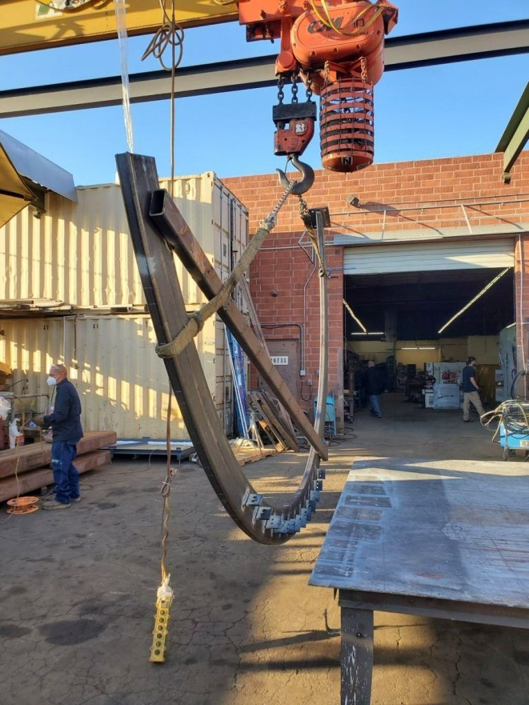 A crane hoists a black metal frame beside a workshop with corrugated siding and open garage doors.