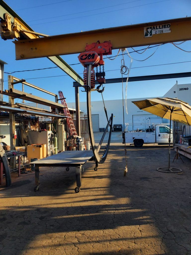 Outdoor industrial yard with a yellow overhead crane, metal worktable, hanging chains, and a white truck by the water.