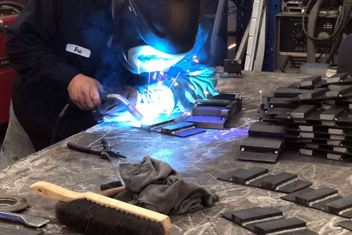 Worker welding metal pieces on a workbench, with bright blue sparks in a workshop.