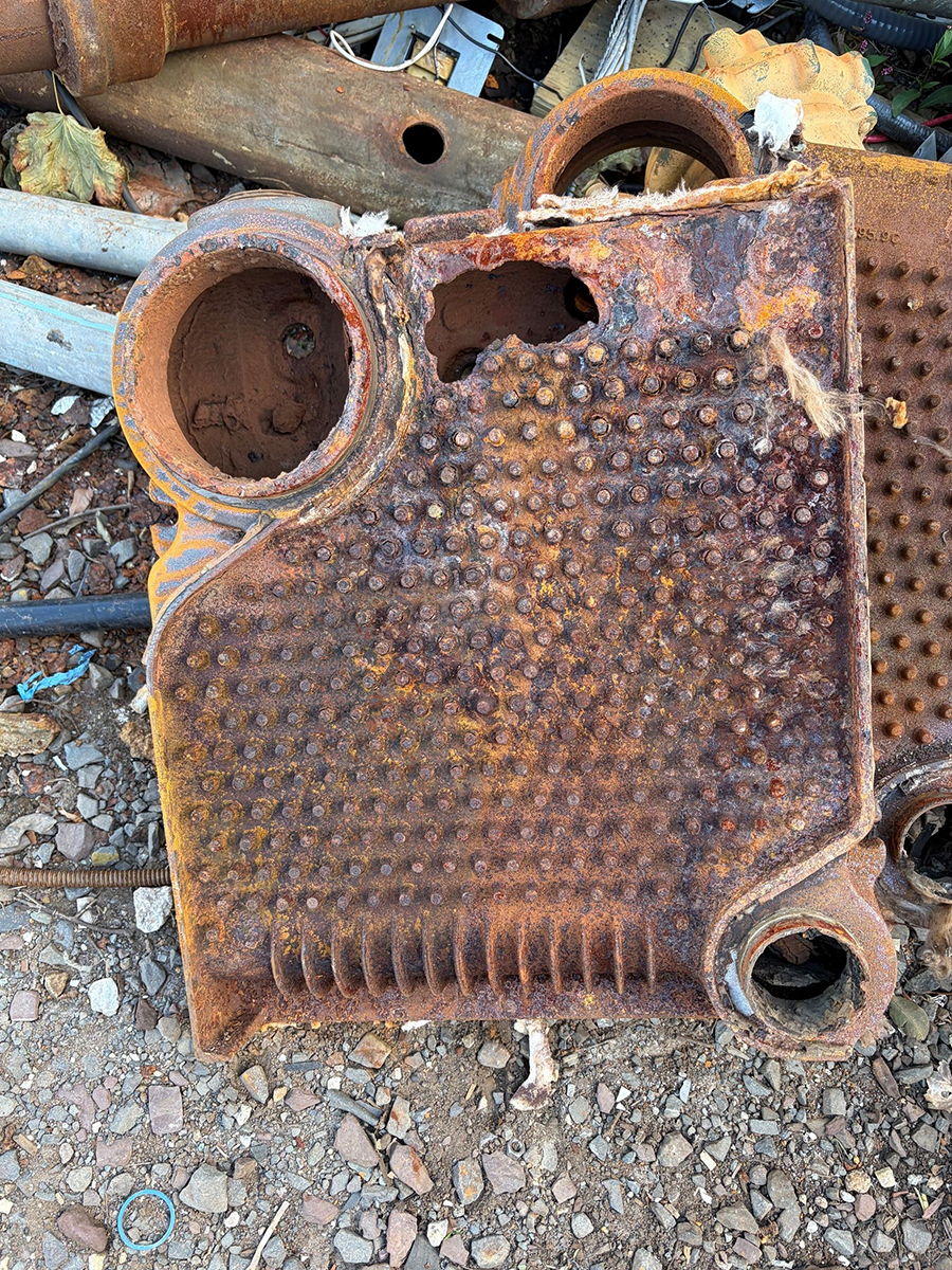 Rusty metal plate with round pipes and a bumpy textured surface lying on gravel and scrap metal.