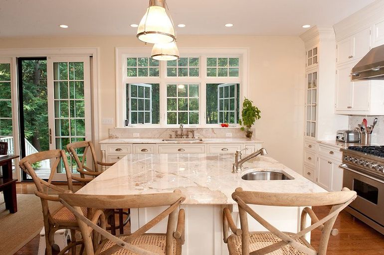Kitchen with island, white cabinets, marble countertop, wooden chairs, and large windows overlooking greenery.