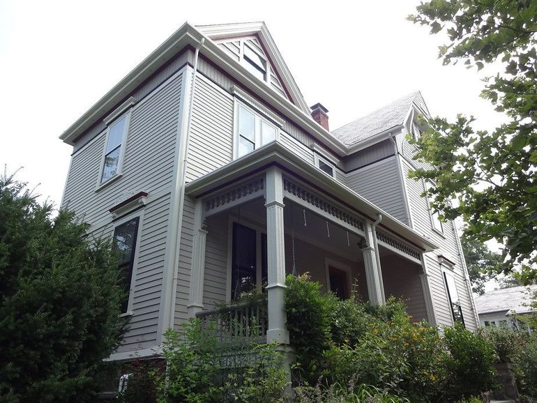 Two-story house with gray siding, white trim, and a front porch, surrounded by green bushes and trees.