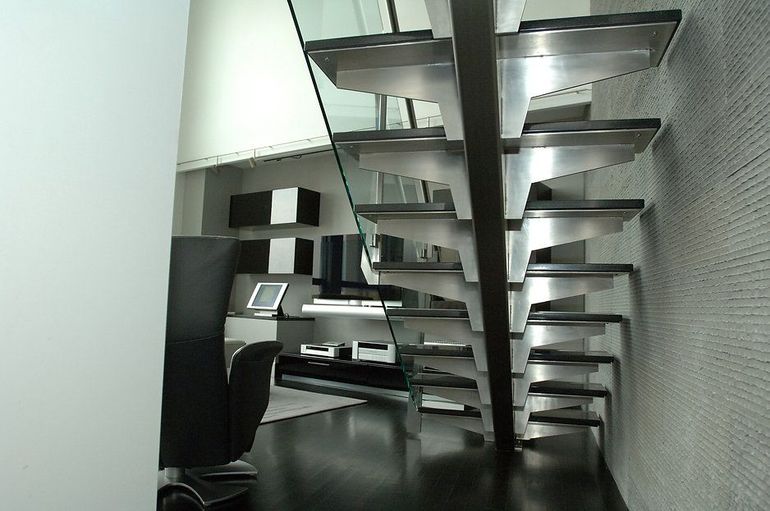 Modern floating staircase with glass railing, leading upward, dark floor, room with black furniture.