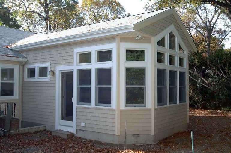 Sunroom addition with multiple windows, beige siding, and white trim against a backdrop of trees and fallen leaves.