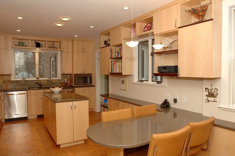 Modern kitchen with light wood cabinets, island, dining table, stainless steel appliances, and window.
