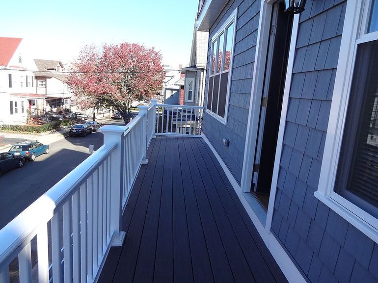 Balcony with dark deck, white railing, and blue siding overlooking a street with cars and buildings.